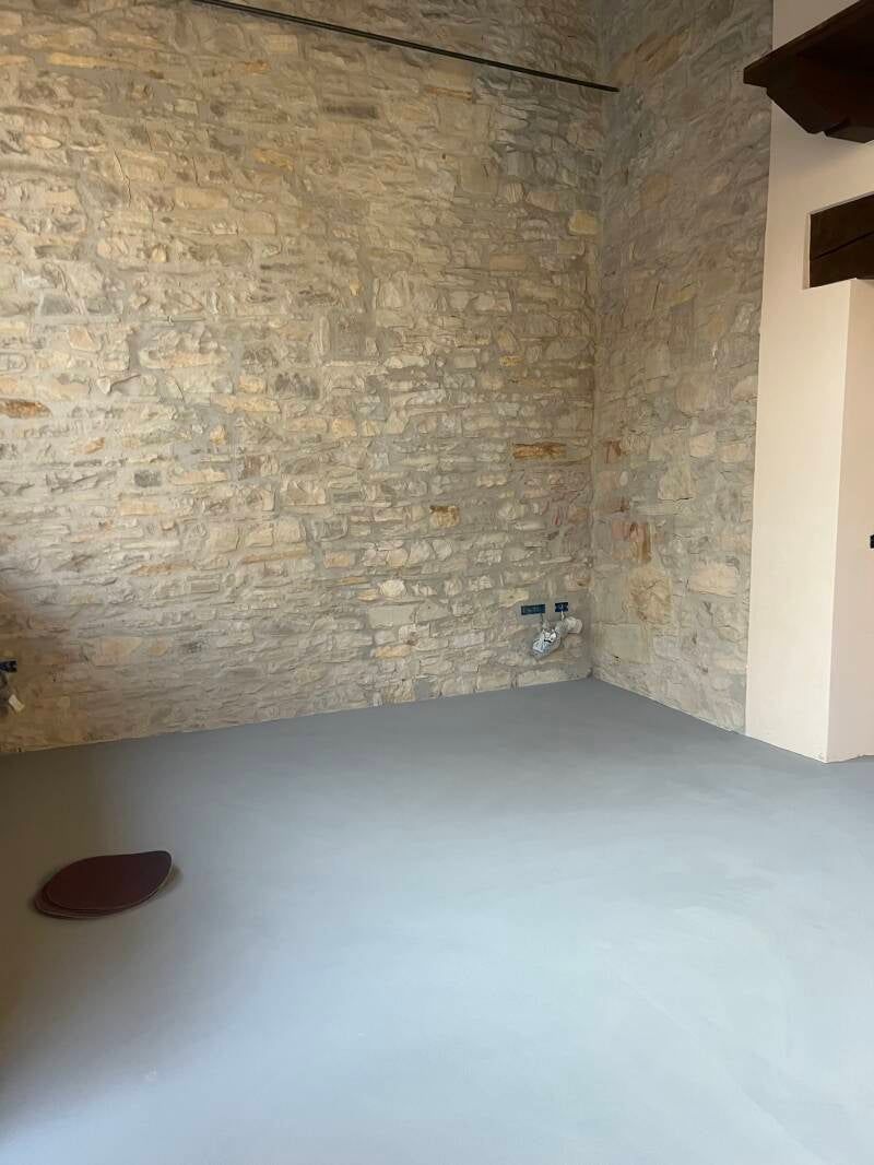 Stanza con pavimento grigio chiaro e pareti in pietra. Impianto idraulico a vista. Travi in ​​legno marrone in alto a destra.