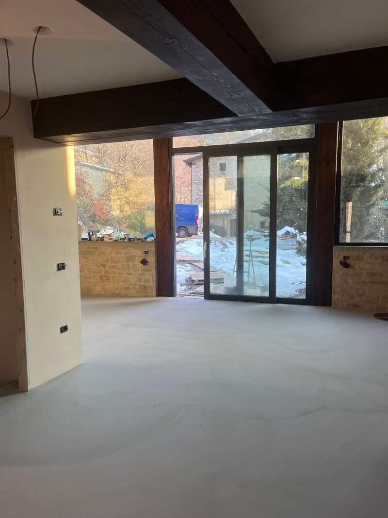 Interno di una stanza con travi in ​​legno a vista, grandi porte in vetro e pareti dai colori chiari, con vista su un paesaggio innevato.