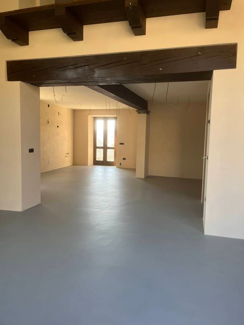 Stanza vuota con pavimento grigio, pareti beige e travi in ​​legno scuro. Porta in fondo.