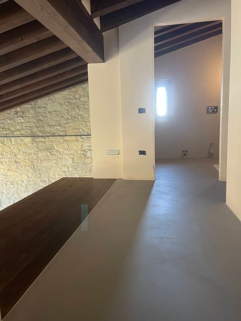 Vista interna di uno spazio ristrutturato con soppalco, muro in pietra, travi in ​​legno e pavimento dipinto di grigio.