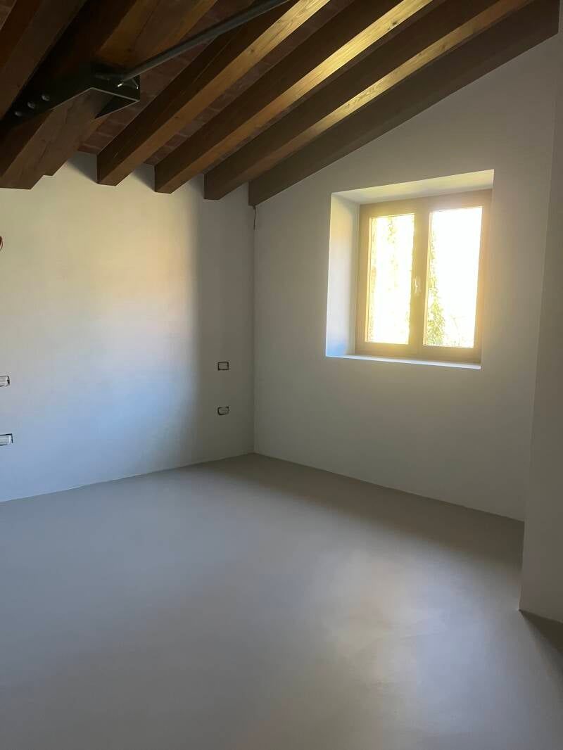 Stanza vuota con pavimento grigio chiaro, pareti bianche, travi in ​​legno e una finestra che lascia entrare la luce del sole.