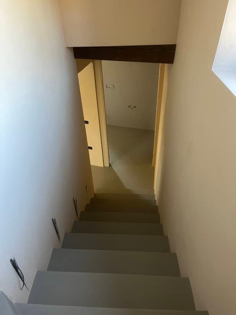Scala che scende e si apre su un corridoio. Gradini grigi, pareti chiare e trave di legno sopra la porta.
