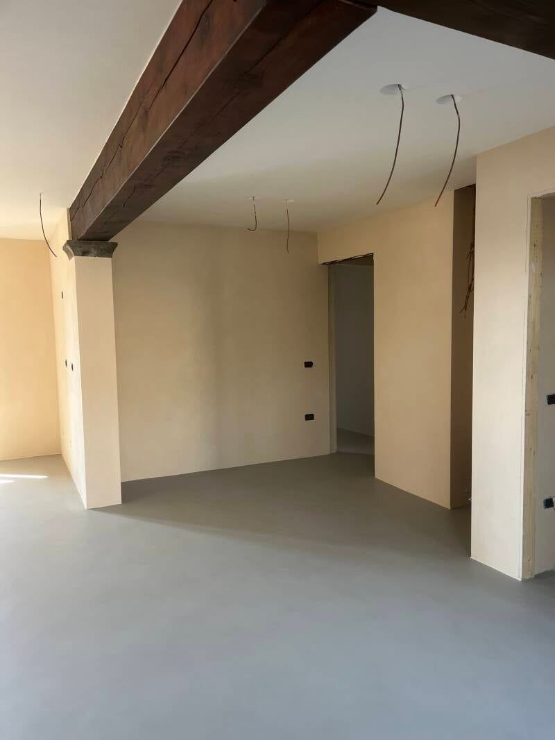 Vista interna di una stanza in costruzione con cavi elettrici a vista, pavimento grigio, pareti beige e una trave in legno scuro.