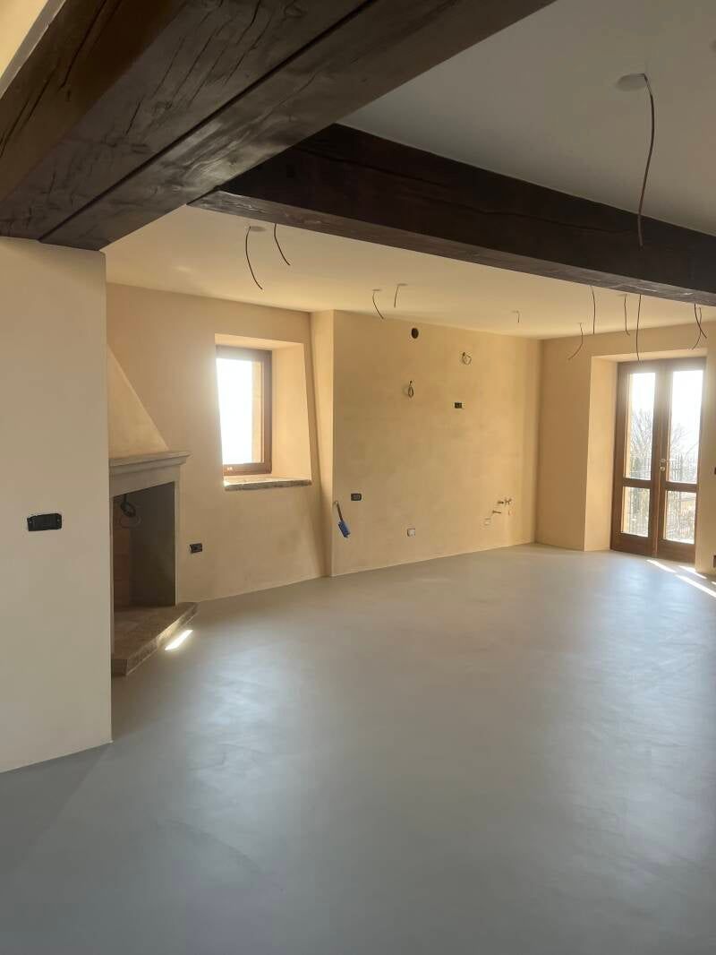 Stanza vuota con pavimento grigio, pareti color cuoio, travi in ​​legno a vista, camino, finestra e doppie porte.