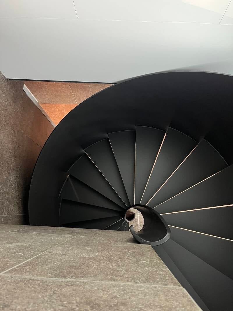 Scala a chiocciola nera su parete e pavimento di colore neutro.