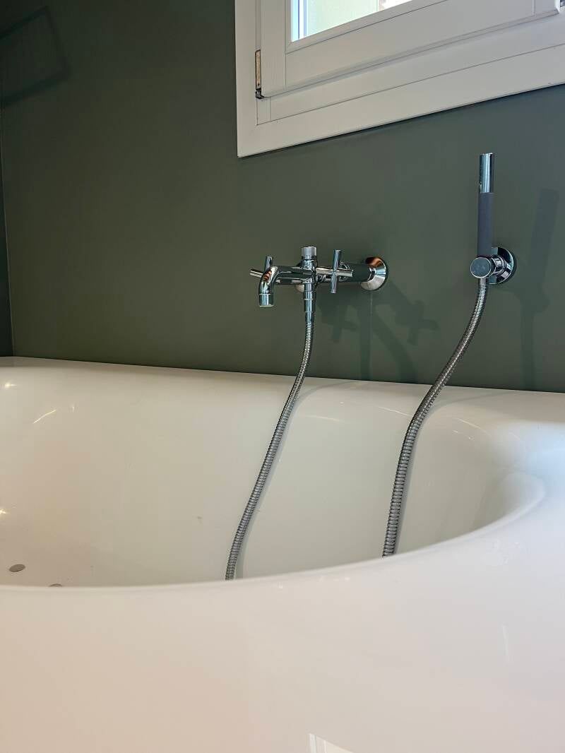 Vasca da bagno bianca con rubinetto argentato e doccetta contro una parete verde, sotto una finestra.