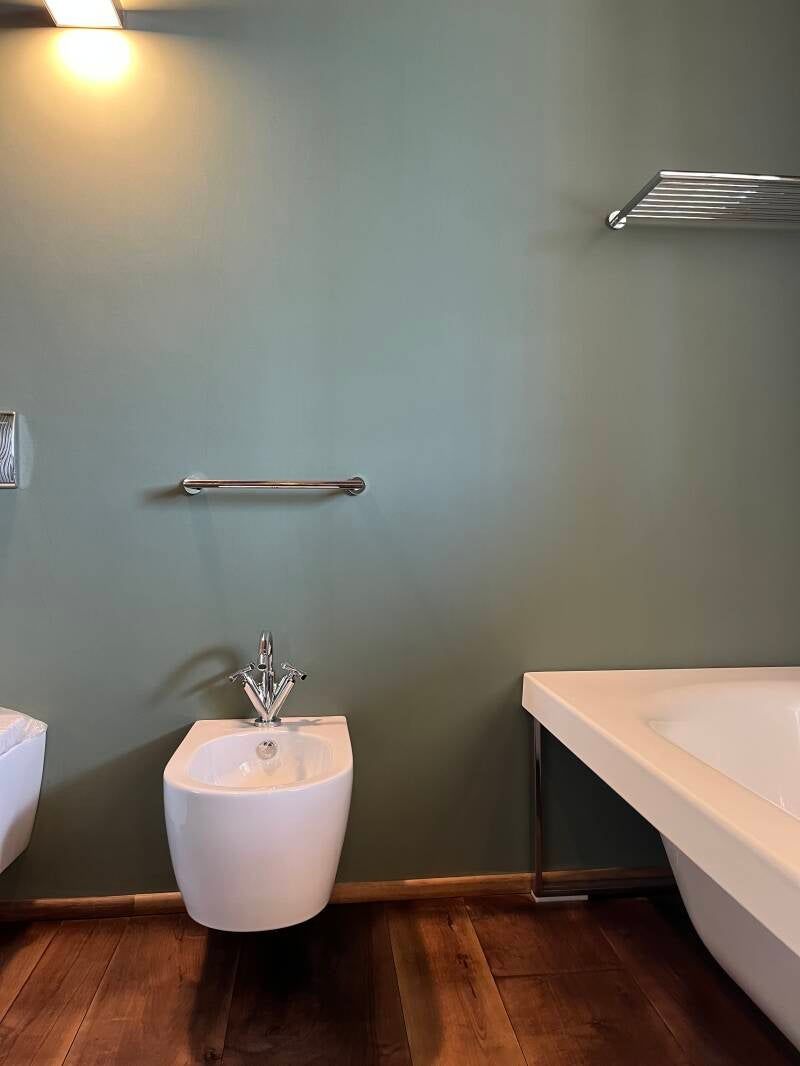 Bagno con bidet bianco, portasciugamani e vasca da bagno contro una parete verde e pavimento in legno.