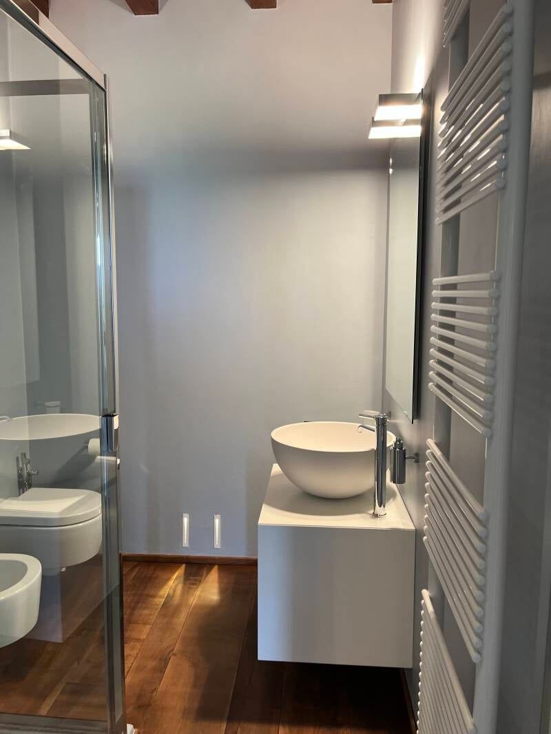 Bagno moderno con lavandino, WC e portasciugamani bianchi; doccia in vetro, pavimento in legno e parete neutra.