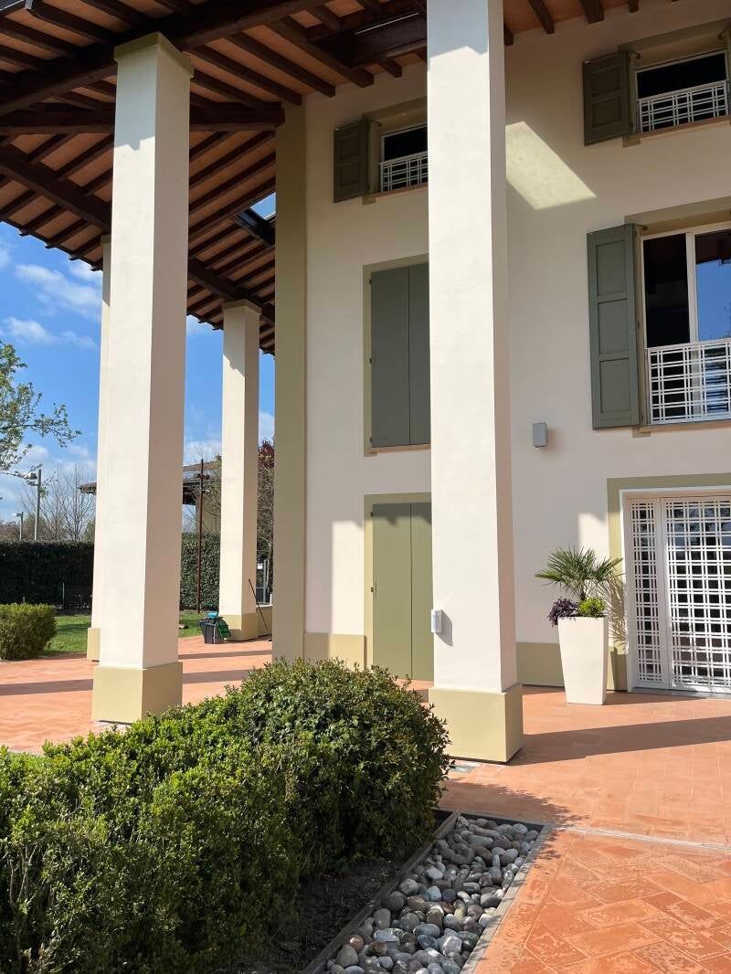 Edificio beige e bianco con alti pilastri e persiane verdi, patio con arbusti.