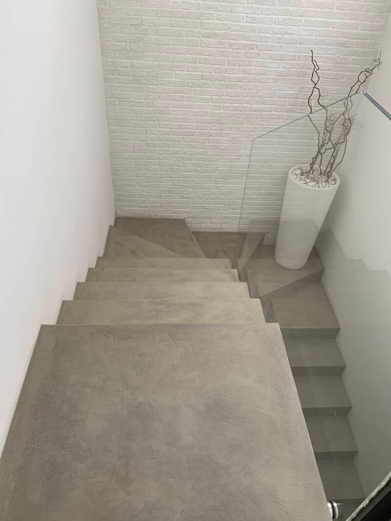 Scala con gradini in piastrelle grigie e parete in mattoni bianchi. Alto vaso bianco con rami sul pianerottolo.