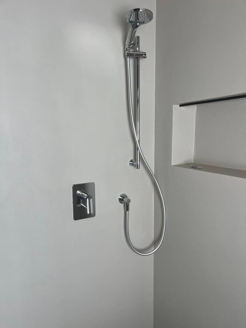 Rubinetto per doccia cromato su parete bianca con mensola incorporata.