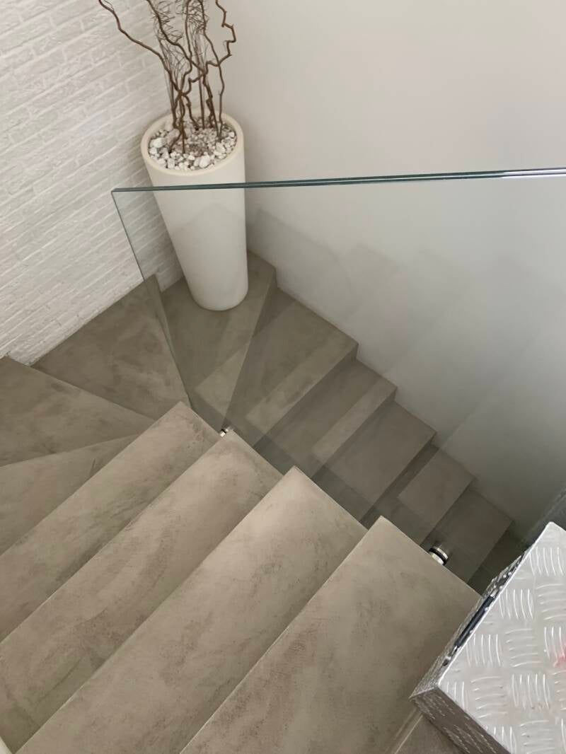 Scala con gradini grigi, ringhiera in vetro e pianta in vaso bianca in un interno moderno.