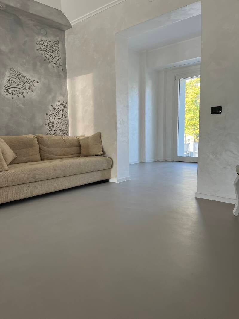 Soggiorno con pavimento grigio, divano beige, parete grigia decorativa e porta luminosa.
