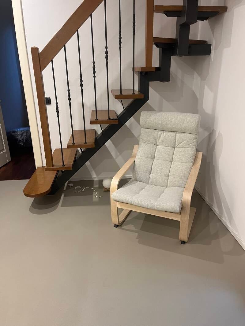 Sedia sotto le scale in legno, cuscino e struttura grigio chiaro, contro una parete e un pavimento grigio chiaro.