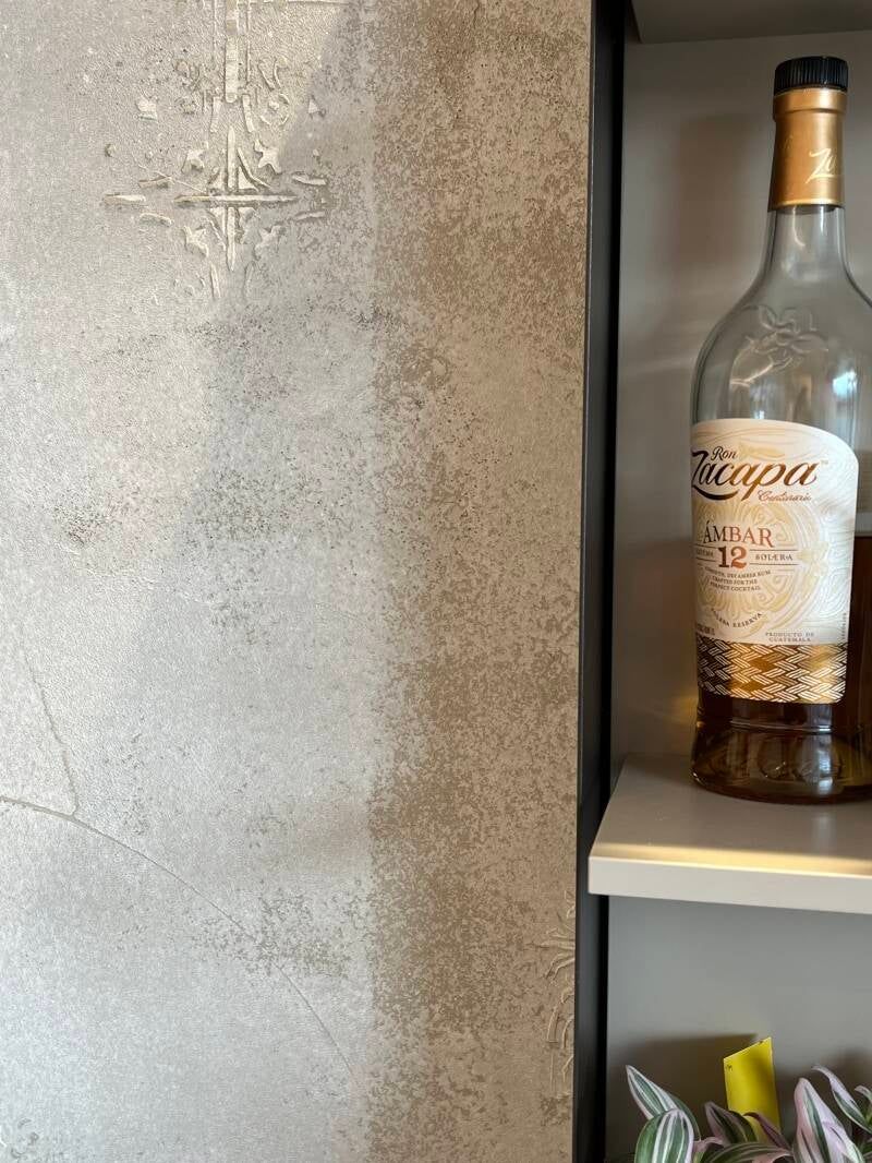 Primo piano di una parete chiara e strutturata accanto a uno scaffale con una bottiglia di liquore.