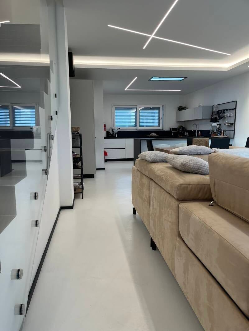 Interni moderni: scale sulla sinistra, soggiorno con divano beige, cucina sullo sfondo, pareti bianche e luci al soffitto.