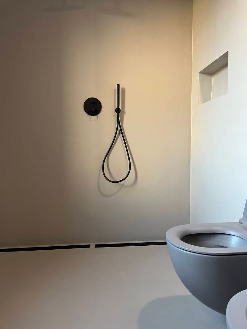 Bagno moderno con WC grigio, soffione doccia nero e pareti e pavimento di colore chiaro.