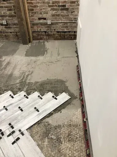 Tile installation in progress: white tiles with spacers on concrete floor, brick and white wall in background.
