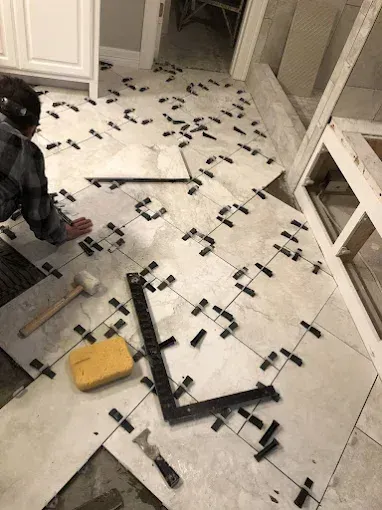 Person tiling a floor with light-colored tiles, using spacers and tools in a room with cabinetry.