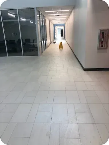 Long hallway with white tiled floor and walls, a caution sign, and windows on the left.