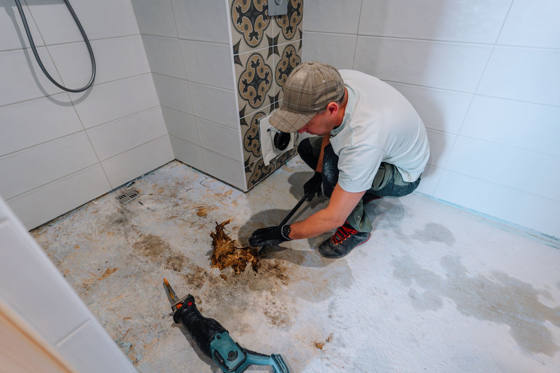 Man in a bathroom using a pry bar and saw to remove flooring.