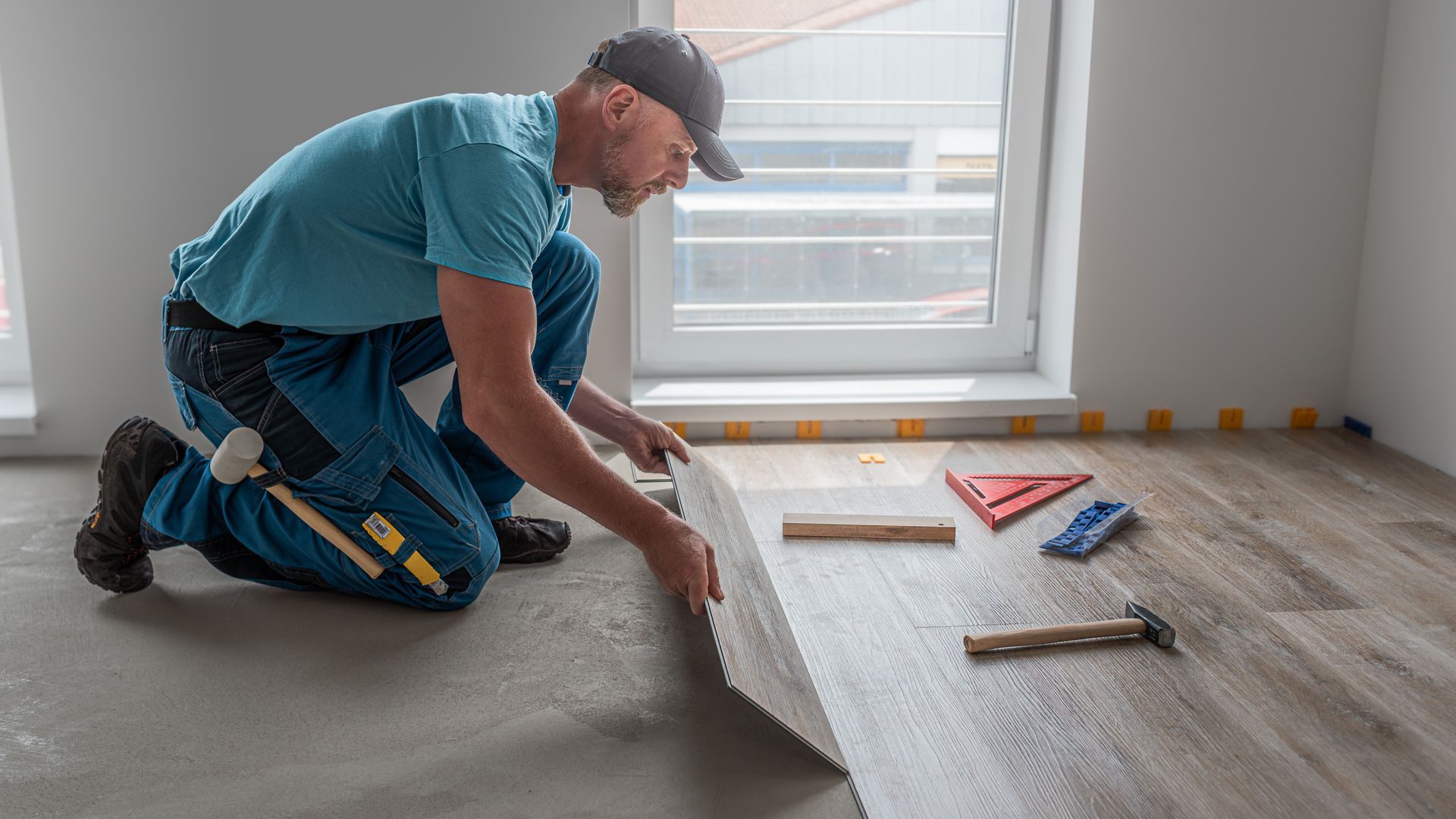 Man installing wood flooring, kneeling in room, holding plank, tools nearby.