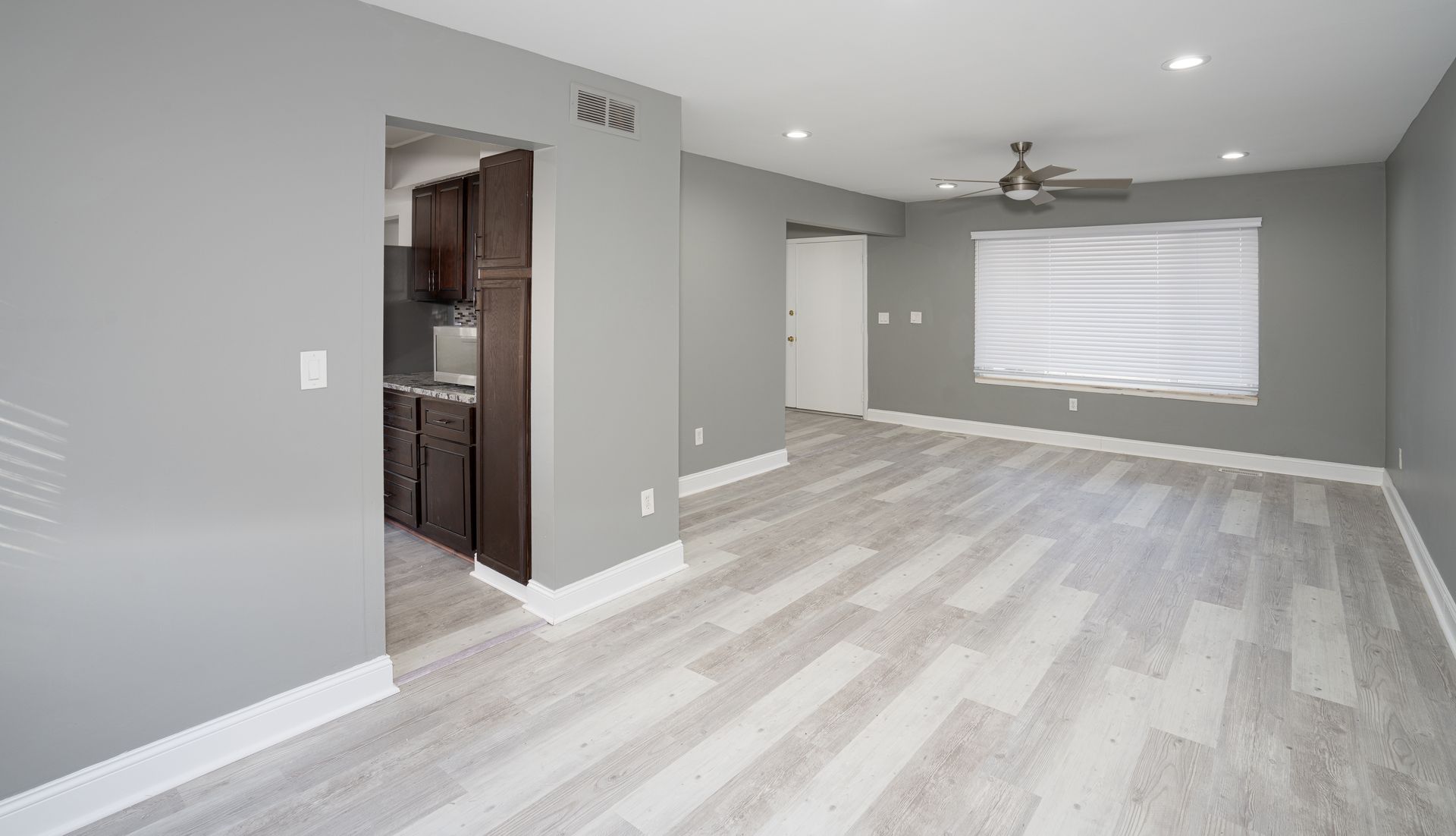 Empty gray-walled living room with gray flooring, white trim, and an open doorway to a kitchen.