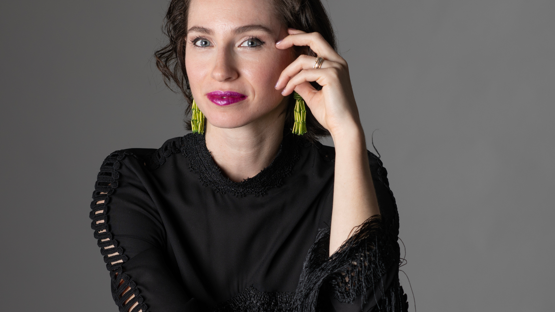 Woman with dark hair, wearing a black top and lime green earrings, smiles with hand near face.