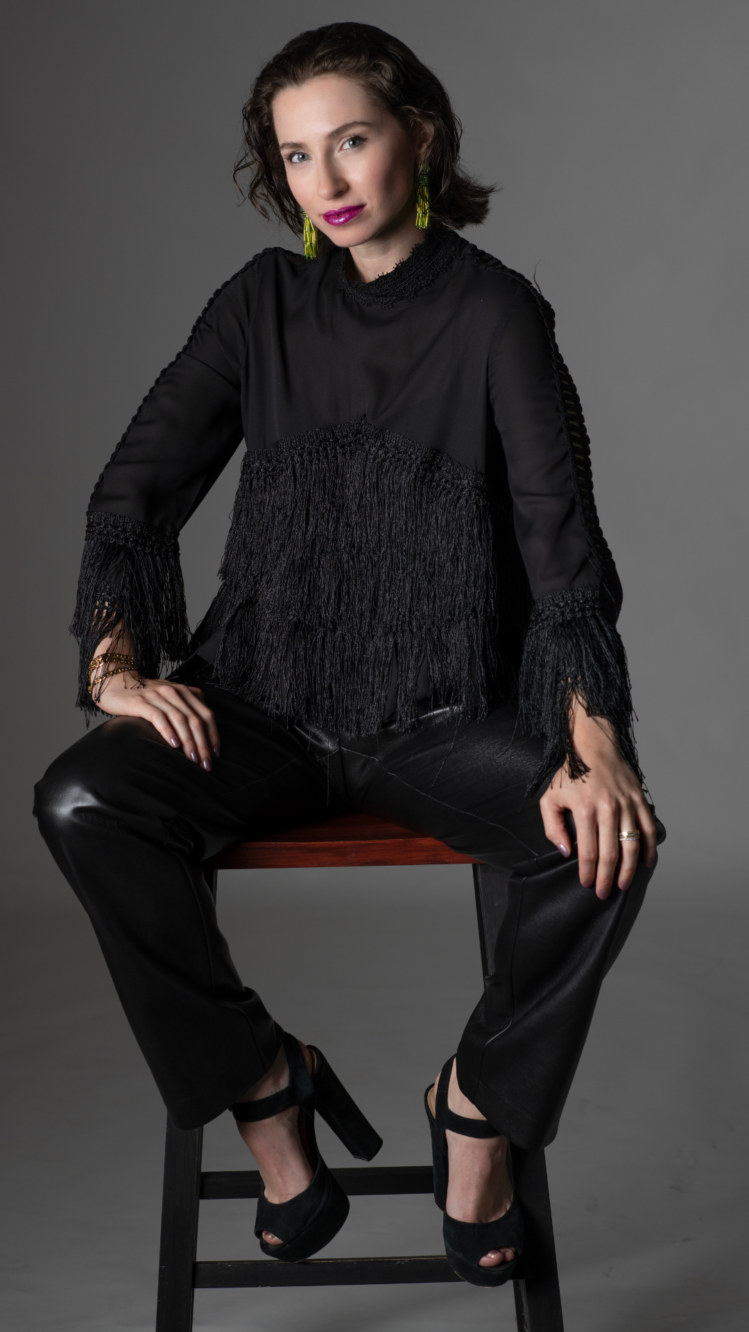 Woman in black top and leather pants sits on a stool, wearing heels.