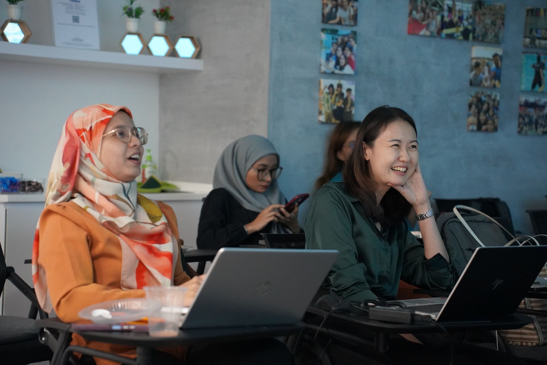 Three women in a classroom, two with laptops, laughing. One wearing a hijab.