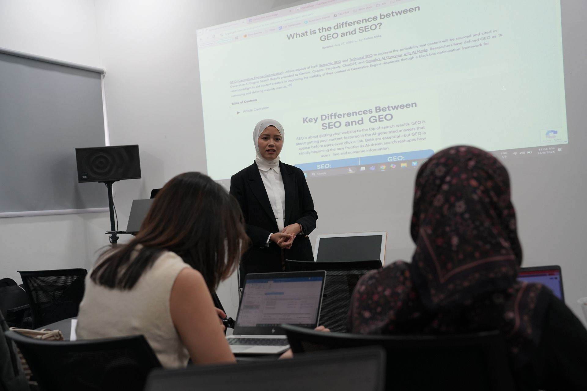 A woman in a hijab presents to a classroom. Students sit at desks with laptops, a projector displays a presentation.