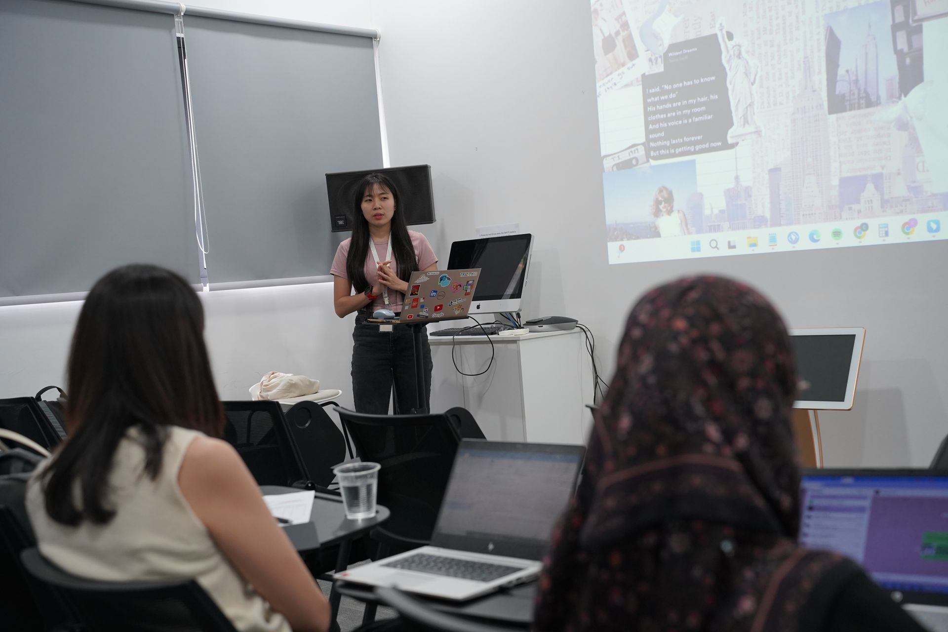 A young woman presents to a small group. She stands in front of a screen with a slideshow.