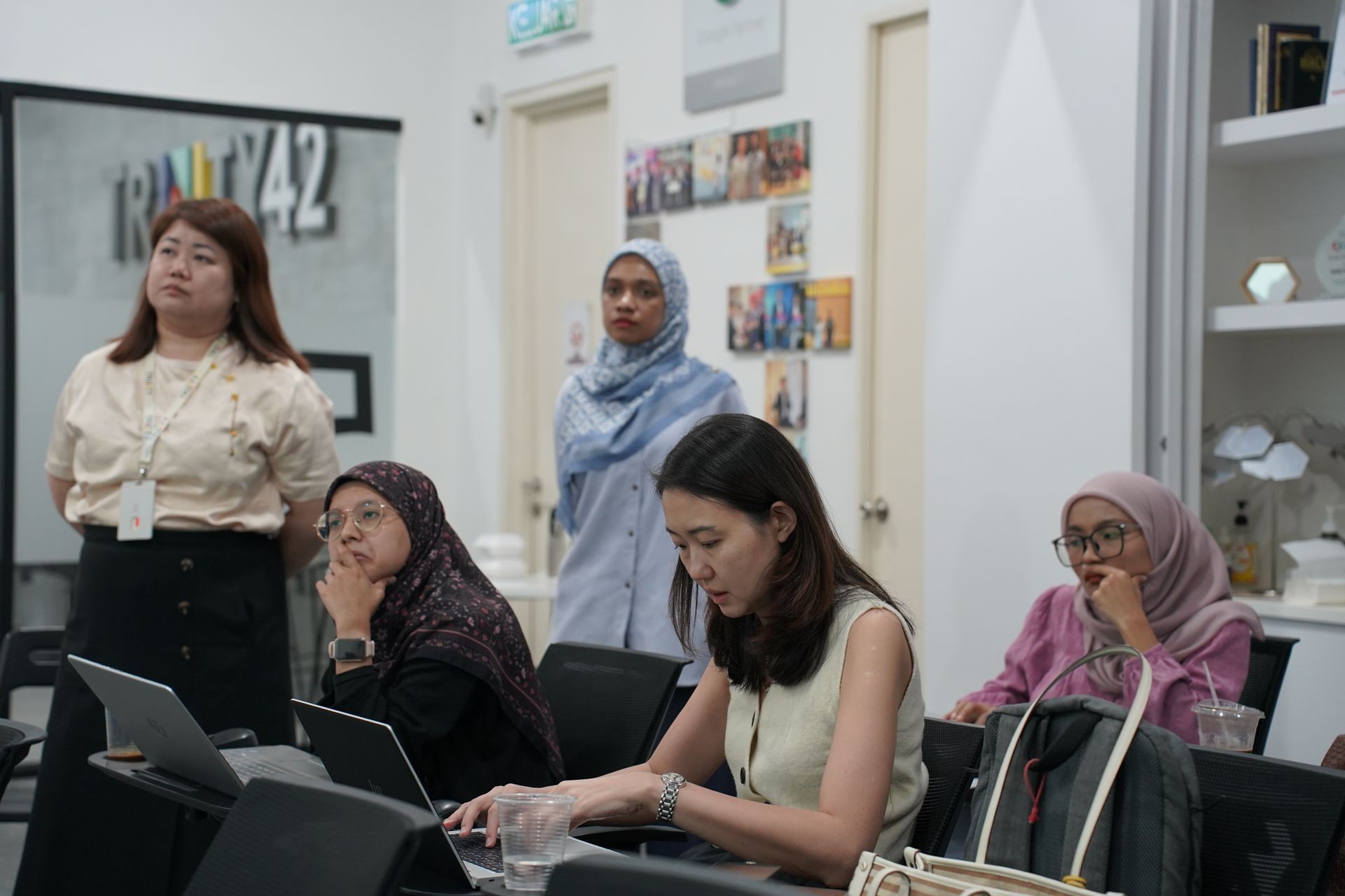 People in an office, some in hijabs, working on laptops, listening, and standing.