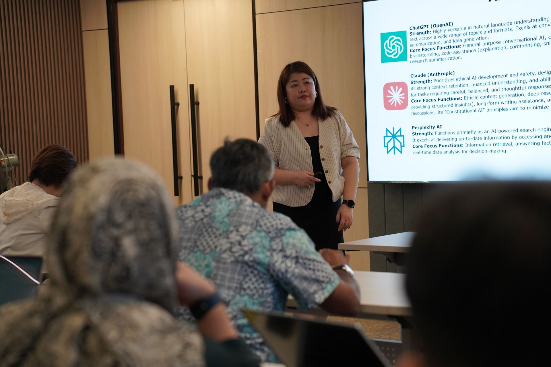 Woman presenting in front of a projector screen to a seated audience.