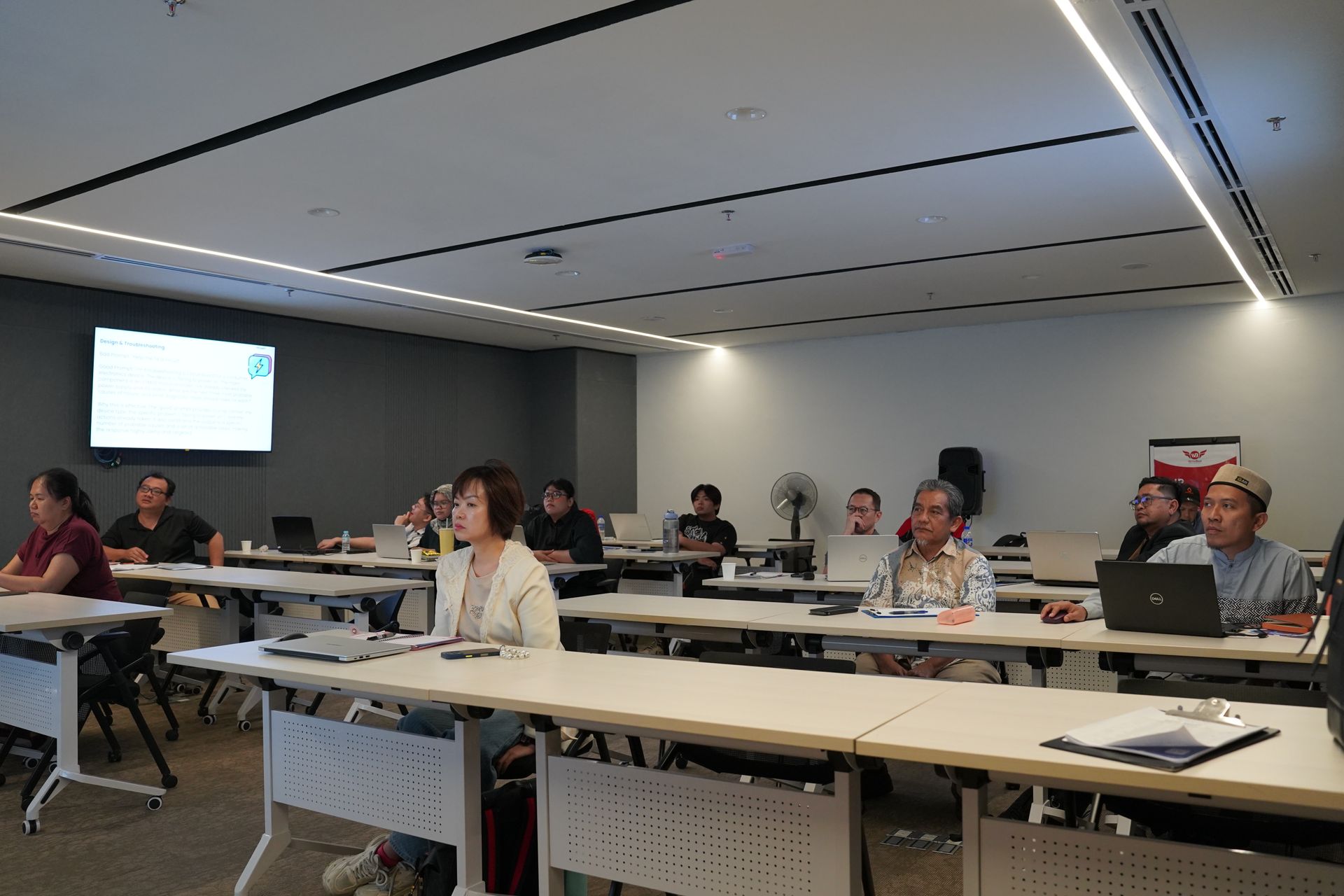 People attending a meeting in a well-lit room, seated at tables with laptops, looking at a screen.