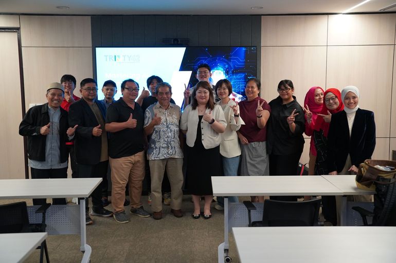 Group of people posing together inside, some giving thumbs up. Background has a screen and tables.