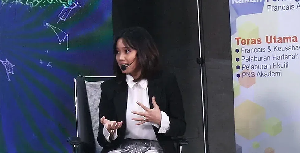 Woman with short hair speaking at a conference, gesturing with hands. Blue and white background, banner on the right.