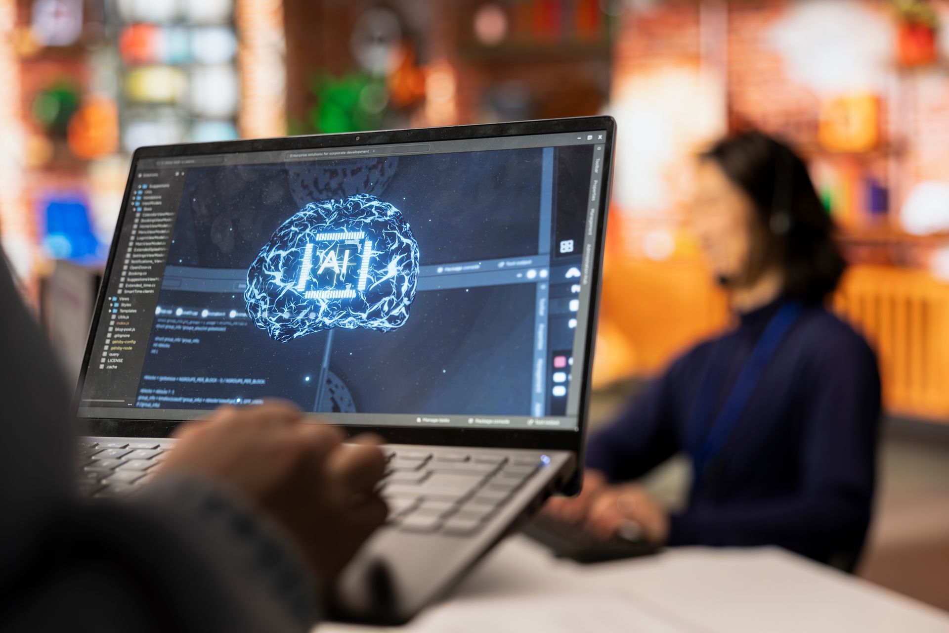 Person coding on laptop, displaying a brain graphic. Another person blurred in background, wearing a headset.