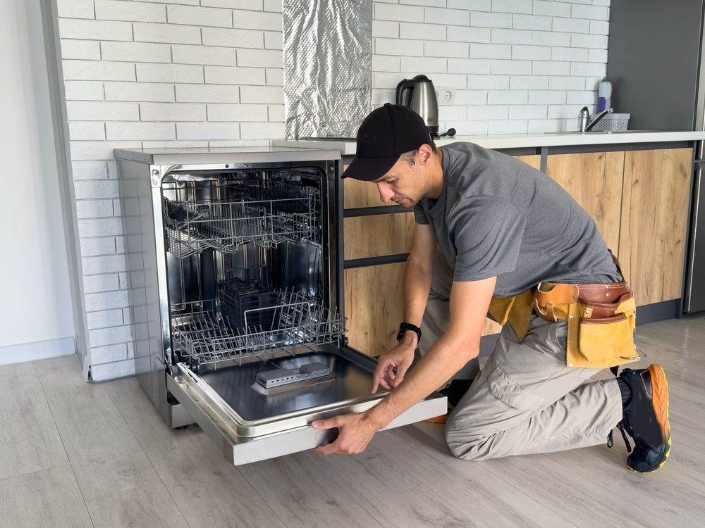 Man kneeling, inspecting dishwasher in a kitchen with exposed cabinets, wearing a tool belt and cap.