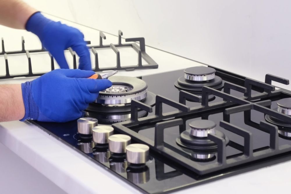 Hands in blue gloves repairing a gas stovetop burner with a tool.