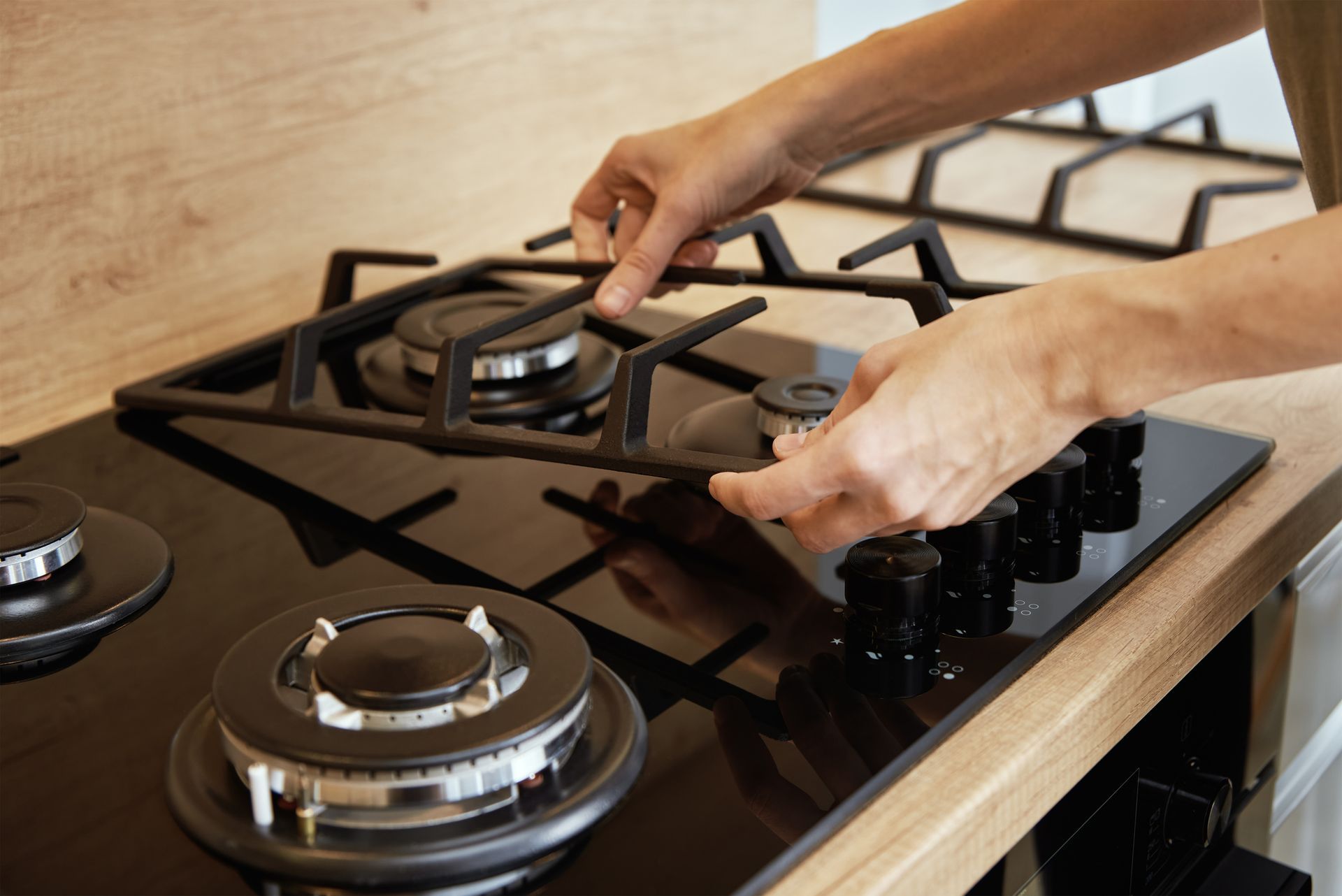 Person placing a black burner grate onto a black gas stove. Wooden countertop visible.