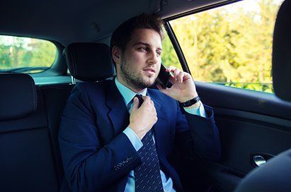 A man in a suit adjusts his tie while on a phone call in the backseat of a car, with a blurred outdoor backdrop.