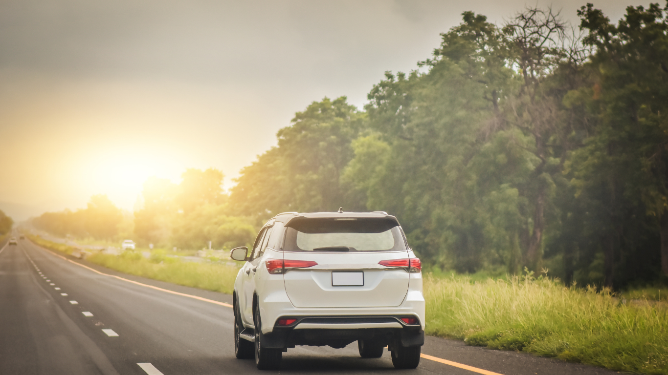 White SUV driving on a highway, sunlit with trees lining the road.