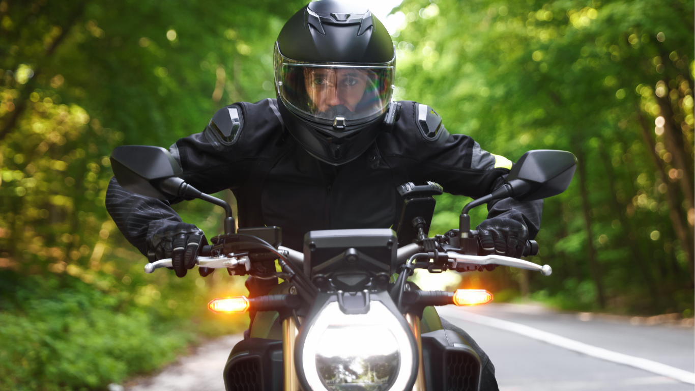 Motorcyclist wearing black helmet and gear riding on a road through a forest.