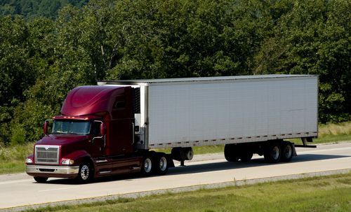 Maroon semi-truck with a white trailer driving on a highway with trees in the background.