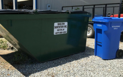 Green dumpster and blue recycling bin on a gravel lot outside a building.