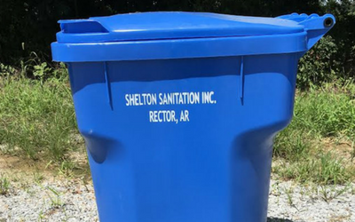 Blue trash bin labeled “Shelton Sanitation Inc., Rector, AR” sitting outdoors on grass
