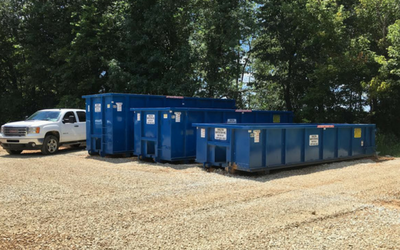 Blue dumpsters and a white pickup truck on a gravel lot beside trees