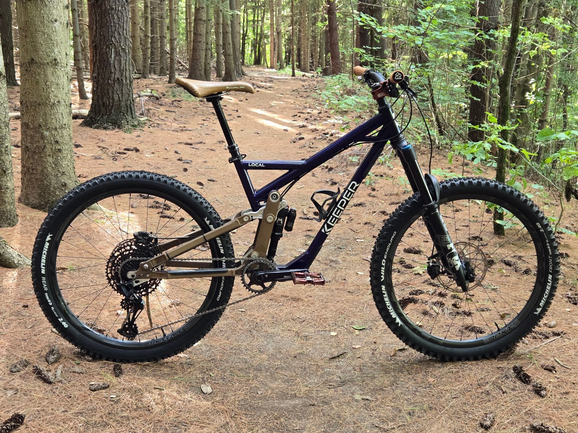 Dark blue mountain bike on a forest trail with brown accents and knobby tires.