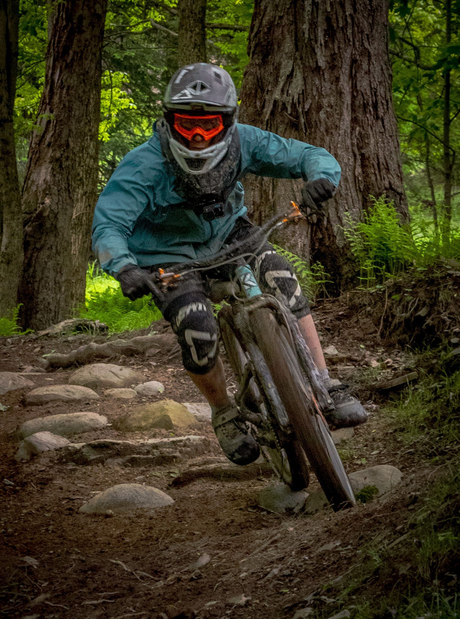 Mountain biker leans into a turn on a rocky forest trail, wearing protective gear.